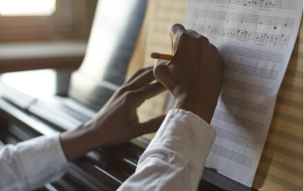 Close-up of dark-skinned hands holding and notating musical paper as it sits on the fall board of a piano.