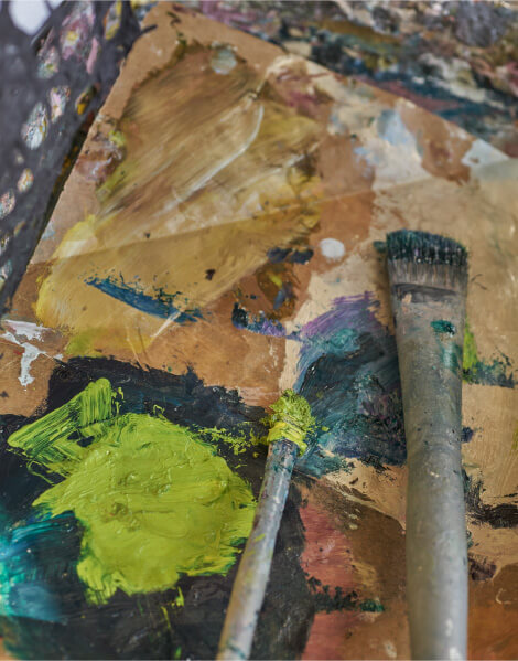 Close-up of a worn paintbrush sitting atop a dirty palette that contains mixes of green, yellow, and brown paint.