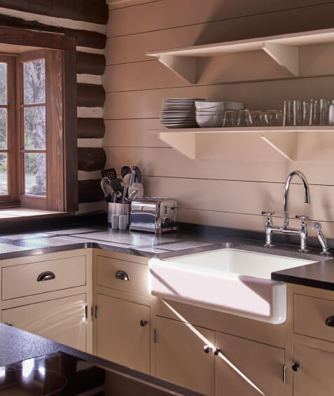 Sunny interior of a cabin kitchen showing exposed log wall, kitchen utensils on shelves, cabinets, granite countertops, and a large ceramic sink.