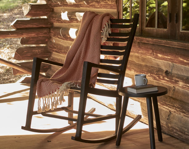 A rocking chair and blanket sit in a sunny location on a cabin porch. To the right there is a book and mug on a side table