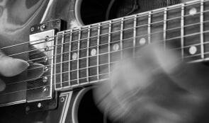 Black and white close-up showing a blurred hand moving along the neck of an electric guitar. A finger from the other hand is cropped near the guitar pickups.