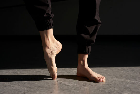 Close-up of lighter-skinned bare feet in black athletic pants. One foot is in a pointe ballet position.