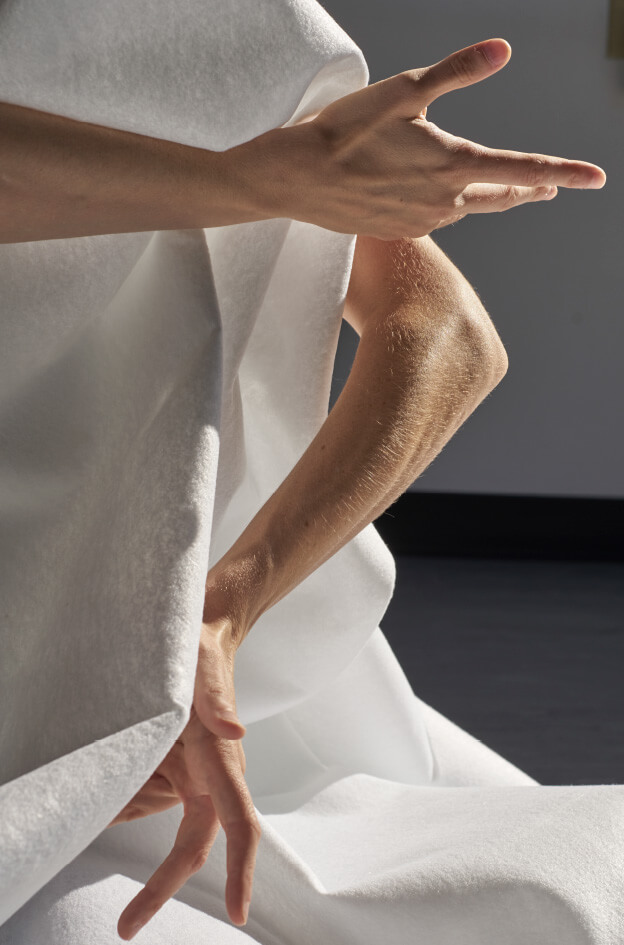 A close up of two white arms interacting with folds of flowing white fabric in a performance setting. One hand gestures to the right, and the other curves downwards. Bright lighting creates dynamic shadows and highlights on both the arms and cloth.