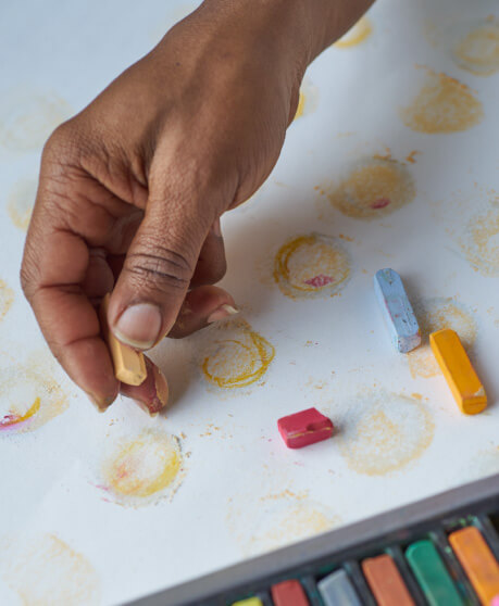 Close-up of a dark-skinned hand using chalk pastel to draw a yellow circle on paper in a larger pattern of predominantly yellow circles. Sky blue, orange, and red pastels of different sizes rest on top of the surface in the bottom right.