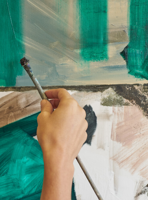 Close-up of a light-skinned hand applying turquoise paint to an abstract painting