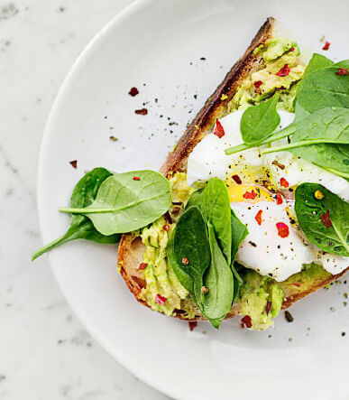 Close-up of avocado toast with a broken poached egg. It is on a white plate on a white marble surface. The plate is garnished with spinach and red pepper flakes