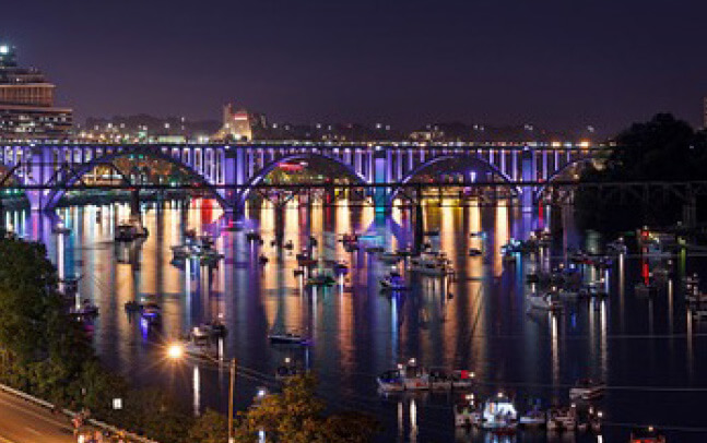 The Henley Bridge at night. The arched cantilever bridge is brightly lit in purple and blue hues. Colored lights reflect on the Tennessee river and many small boats populate the water.