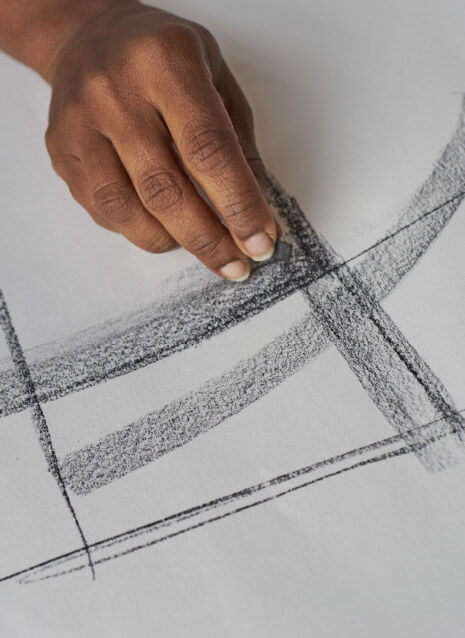 Close-up of a cropped, dark-skinned hand drawing a thick arching line with the side of a piece of charcoal over a piece of paper.