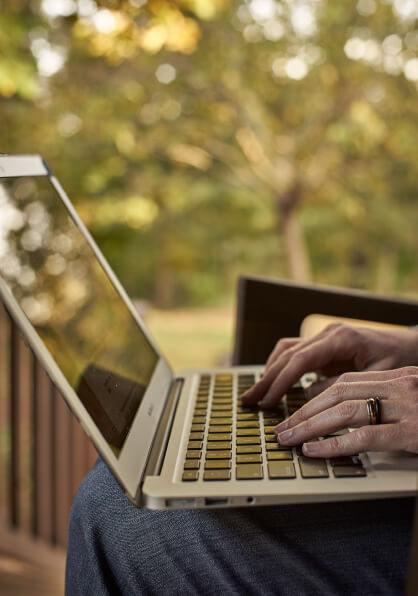 Shallow-focus, cropped close-up of a light-skinned woman's hands using a laptop keyboard on an outdoor porch. Out-of-focus trees exist in the background.