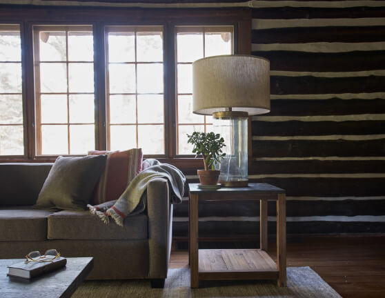 A row of windows built into a rustic log cabin wall illuminates a living room with a couch with pillows and blankets. Adjacent to the couch is a side table with a glass lamp and a small potted plant.