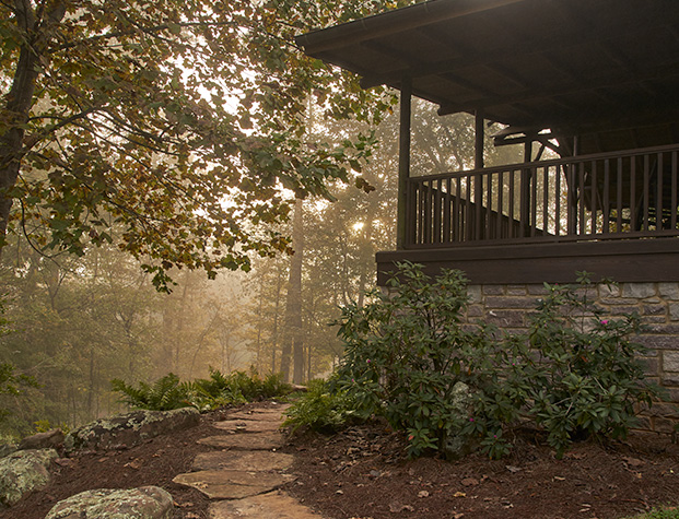 A stone path recedes around the corner of a covered cabin patio on a foggy morning.