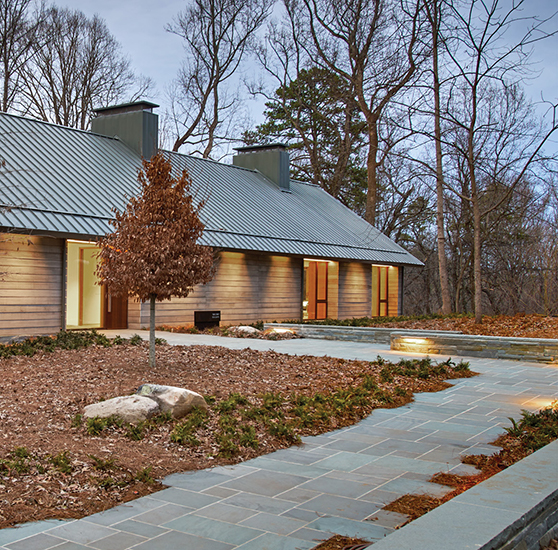 A lit stone path at dusk leads up to the McDonough House on the left, the main building of Loghaven. Two large chimneys can be seen on a sloping metal roof, and the exterior walls are wood-paneled.