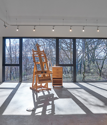 A large wooden easel and taboret sit in front of floor to ceiling windows overlooking a wooded drop-off in the fine art studio. Track lights can be seen lining the ceiling above the windows.