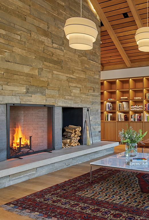 A lit fire and firewood inside of a large stone fireplace in the McDonough House living room. A portion of the Loghaven Library can be seen on wooden shelves to the right, and a white marble coffee table sits in front of the fire.