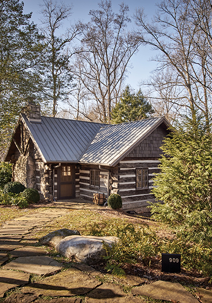 Exterior of a cabin at Loghaven. Surrounded by trees and shown in daylight, the cabin walls are made up of dark logs and white mortar. The large chimney is cobblestone, and the roof is gray metal.