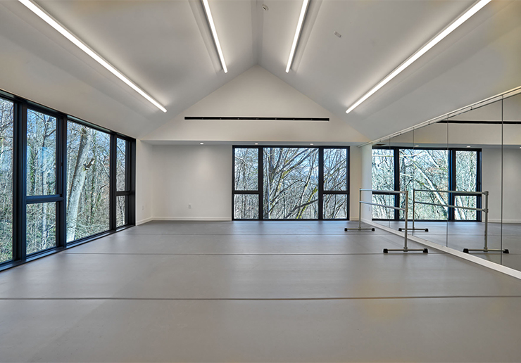 The interior of Performing Arts Studio is pictured with a grey marley floor, two white walls, and one mirrored wall. The windows look out on trees and blue sky.