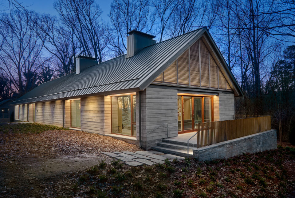 The McDonough House pictured at night. A patio is to the right, and the exterior is a combination of wood panel and floor to ceiling windows. The pitched metal roof is gray, and two large chimneys can be seen. 